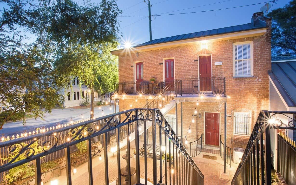 A two-story brick building with a balcony, a staircase with decorative railings, and string lights.