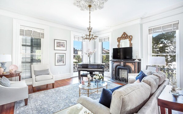 A spacious living room with a grand piano, a fireplace, and a large window offering a view of the outdoors.