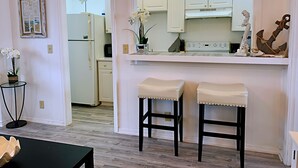 A kitchen with white cabinets, a white refrigerator, and a white stove. There are two stools with white cushions and black legs. A small table with a vase of flowers is visible.