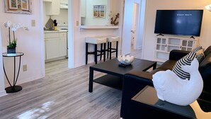 A modern living room with a black sofa, a coffee table, and a TV. There is a kitchen in the background with white cabinets and a dining area with a white table and chairs.