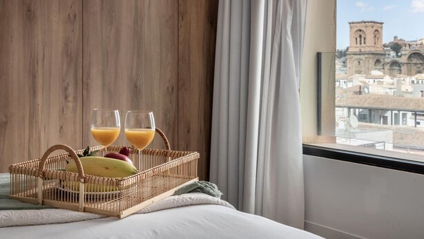 Habitación de hotel con pared de madera, ventana con vistas a un edificio histórico, cama con ropa blanca y cesta de mimbre con dos vasos de jugo de naranja.