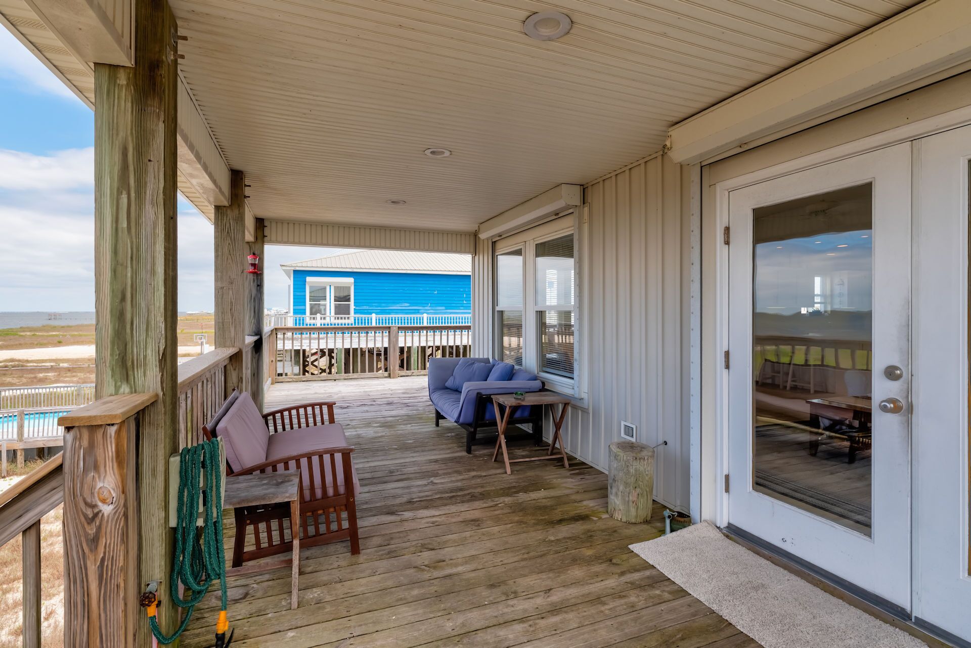 A covered porch with a wooden deck, a seating area with a table and chairs, and a view of a blue building across the beach.