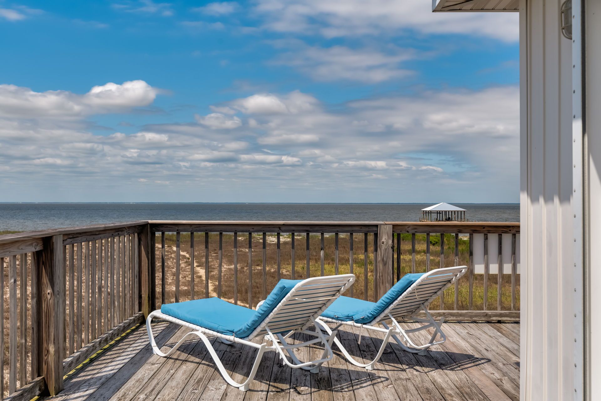 Une terrasse en bois avec deux chaises longues blanches dotées de coussins bleus, offrant une vue sur une plage et l’océan.