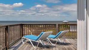 A wooden deck with two white lounge chairs with blue cushions, overlooking a beach and the ocean.