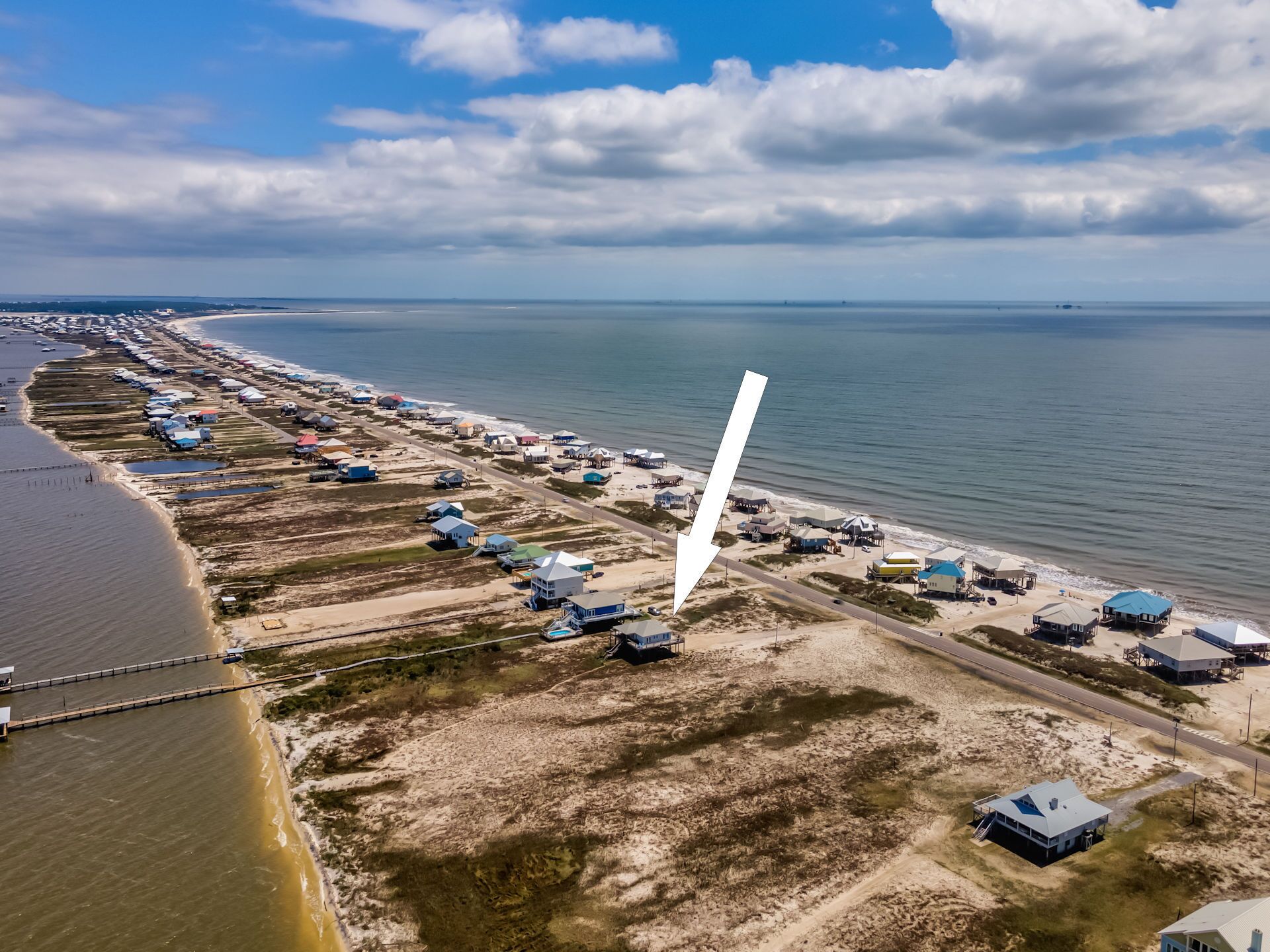 Vue aérienne d’une zone côtière avec une longue rangée de maisons de plage le long du rivage, une route qui longe la plage et une vaste étendue d’eau en arrière-plan.