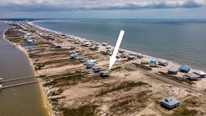 An aerial view of a coastal area with a long row of beach houses along the shoreline, a road running parallel to the beach, and a large body of water in the background.