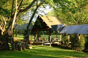 Een rustieke houten tuinpaviljoen met rieten dak, omgeven door een verzorgd gazon en bomen.
