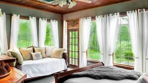 Un dormitorio con dos camas, cabecera de madera, un escritorio con silla y ventanas grandes con cortinas blancas.