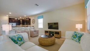 Una sala de estar moderna con sofás blancos, una mesa de centro y un televisor. Se observa una cocina al fondo con armarios oscuros y un microondas.