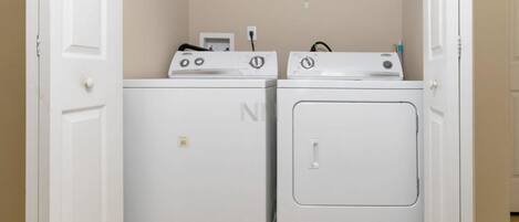 A laundry room with two white washing machines and a dryer, a shelf above the machines, and a door on the left.