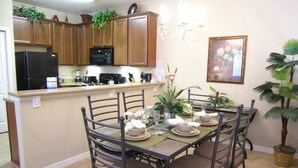A dining area with a round table set for four, surrounded by metal chairs. There is a kitchen area with wooden cabinets and a refrigerator in the background.