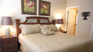 A neatly made bed with a beige bedspread and pillows, flanked by two bedside tables with lamps, in a room with a door and a wall-mounted decorative item.