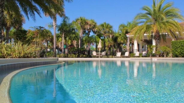 A clear swimming pool with a curved edge, surrounded by palm trees and a building with columns. - Oakwaterresort Near Disney h (St. Cloud)