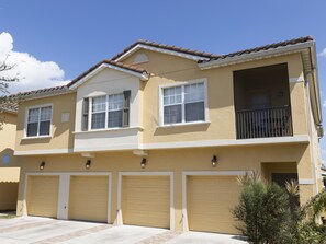 Il s’agit d’un immeuble résidentiel à deux étages, doté d’une façade jaune, de bordures blanches et d’un toit recouvert de tuiles brunes. On y trouve trois portes de garage, un balcon à l’étage supérieur et un petit arbre dans le jardin avant.