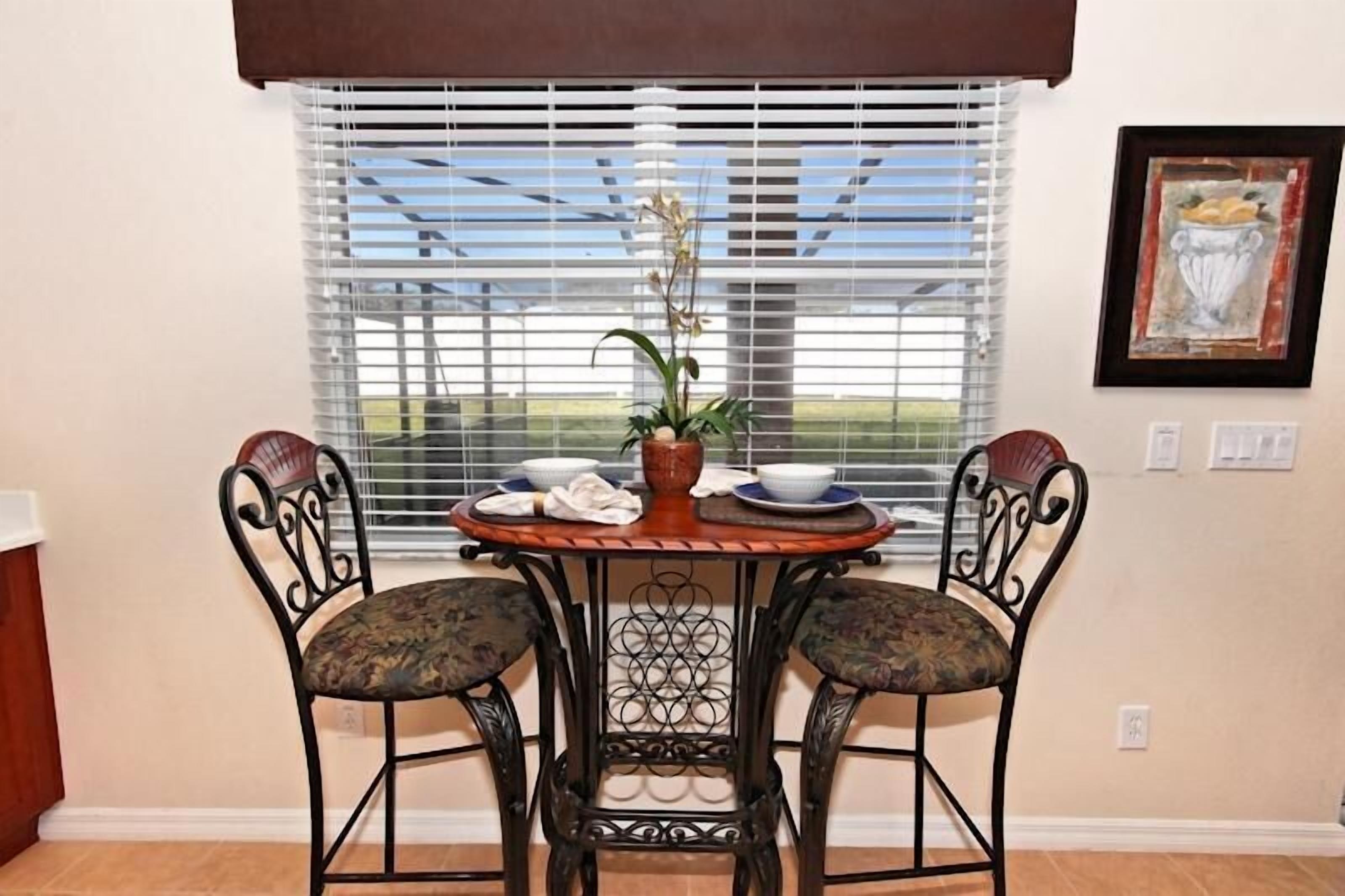 A dining area with a round table, two chairs, a potted plant, and a window with blinds.