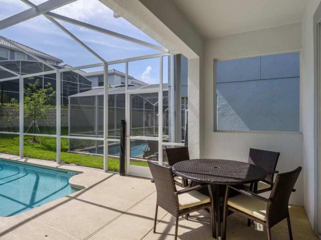 A covered outdoor area with a pool, a table with chairs, and a view of a house through a glass enclosure.