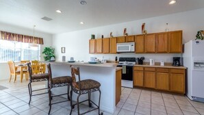 A kitchen with wooden cabinets, a white refrigerator, a microwave, and a dining area with two chairs. - 113bll Disney 5 Bedroom Pool Home With Games Room (Bartow)
