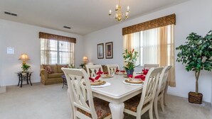 A dining area with a white table set for six, surrounded by white chairs. There are two windows with curtains, a chandelier, and a potted plant. - 113bll Disney 5 Bedroom Pool Home With Games Room (Bartow)