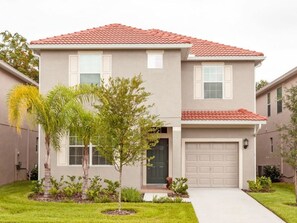A two-story house with a red-tiled roof, a front garage, and a small front yard with palm trees and shrubs. - Paradise Palms 6 Bedrooms 5 Baths Best (Kissimmee)