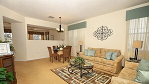 A living room with a beige sofa, a glass coffee table, and a patterned rug. There is a dining area with a table and chairs in the background. - 103bll Fantastic 4 bed With Games Room (Bartow)