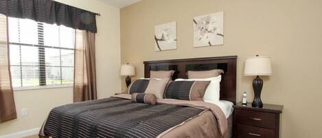 A bedroom with a large bed, two bedside tables, a window with blinds, and two wall-mounted artworks.