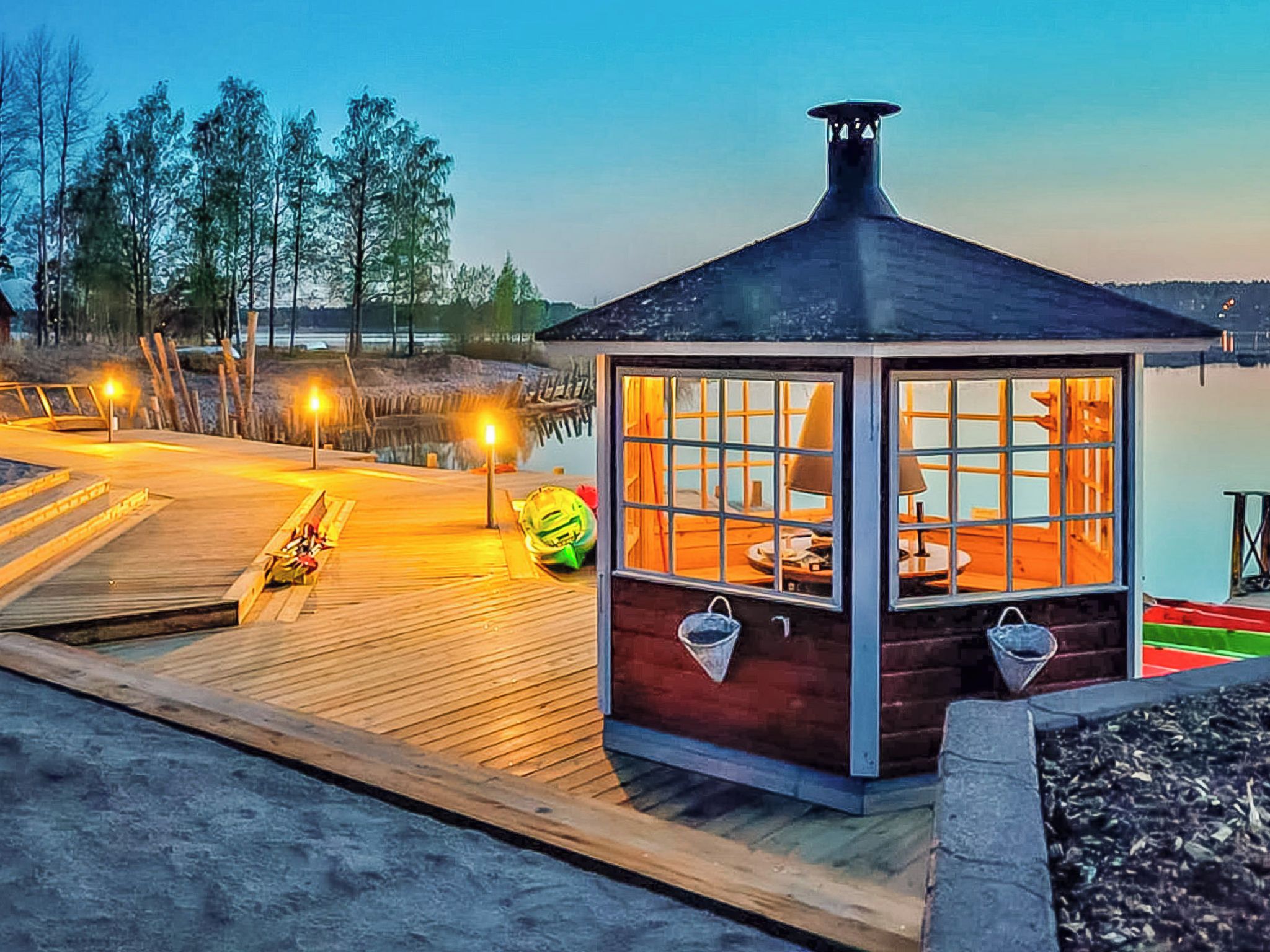 A wooden deck leading to a small, dark-roofed structure with windows and a chimney, overlooking a body of water and trees.