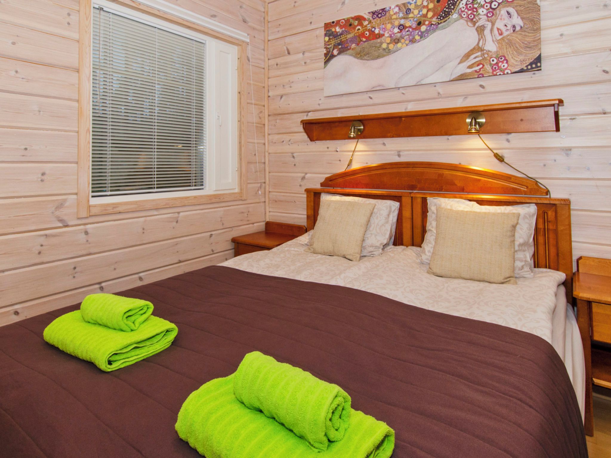 Ein Zimmer mit Holzwänden, einem Bett, zwei Kissen, einem Nachttisch, einer Lampe und einem Fenster mit Jalousien.