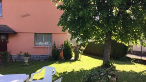 A garden area with a white plastic table and chairs, a small tree, and a building with a pink facade. - Maison Corisande, near the entrance to Bagnères de Bigorre (Gerde)