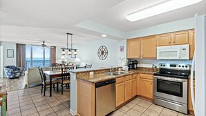 A modern kitchen with wooden cabinets, a stainless steel stove, and a dishwasher. There is a dining area with a table and chairs, and a large window overlooking the ocean. - Updated Oceanfront Condo, Indoor and Outdoor Pools | Crescent Shores - N 303 (North Myrtle Beach)