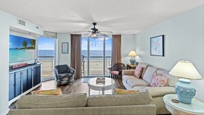 A spacious living room with a large TV, comfortable seating, a ceiling fan, and a view of the ocean. - Updated Oceanfront Condo, Indoor and Outdoor Pools | Crescent Shores - N 303 (North Myrtle Beach)