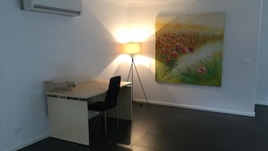 A small office space with a desk, chair, and a lamp. There is a painting on the wall and an air conditioning unit mounted on the wall. - AJ's Home. Modern, open plan & quiet location. (Sale)