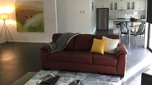 A living room with a red sofa, a coffee table with books, a painting on the wall, and a view into the kitchen. - AJ's Home. Modern, open plan & quiet location. (Sale)