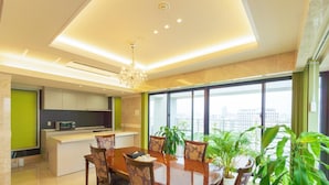 A modern dining area with a large table, chairs, and a chandelier. There is a kitchen area with cabinets and a microwave. The room has large windows with a view of the city.