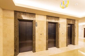 A hallway with four elevators, each with a metallic door and a small sign above. The walls are tiled in a light beige color, and the floor is polished. - Hotel Lantana Naha Kokusai - Dori (Naha)