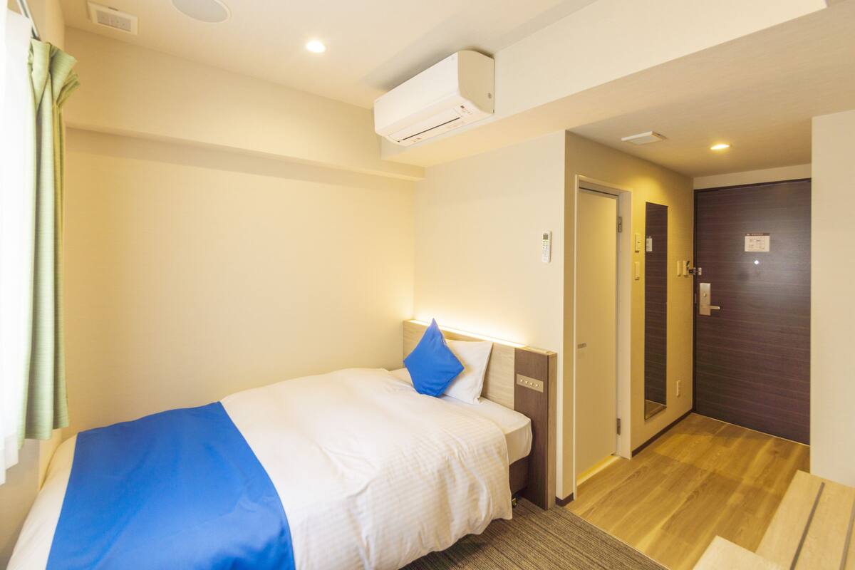 A hotel room with a bed, an air conditioner, a door, and a wooden floor.