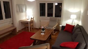 Un salon avec une table en bois, un canapé gris, un tapis rouge et une fenêtre avec des rideaux.