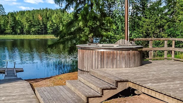 A wooden deck with a hot tub and a dock extending into a lake, surrounded by dense forest.