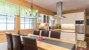 A wooden interior with a dining table, chairs, a chandelier, and a kitchen area with appliances.