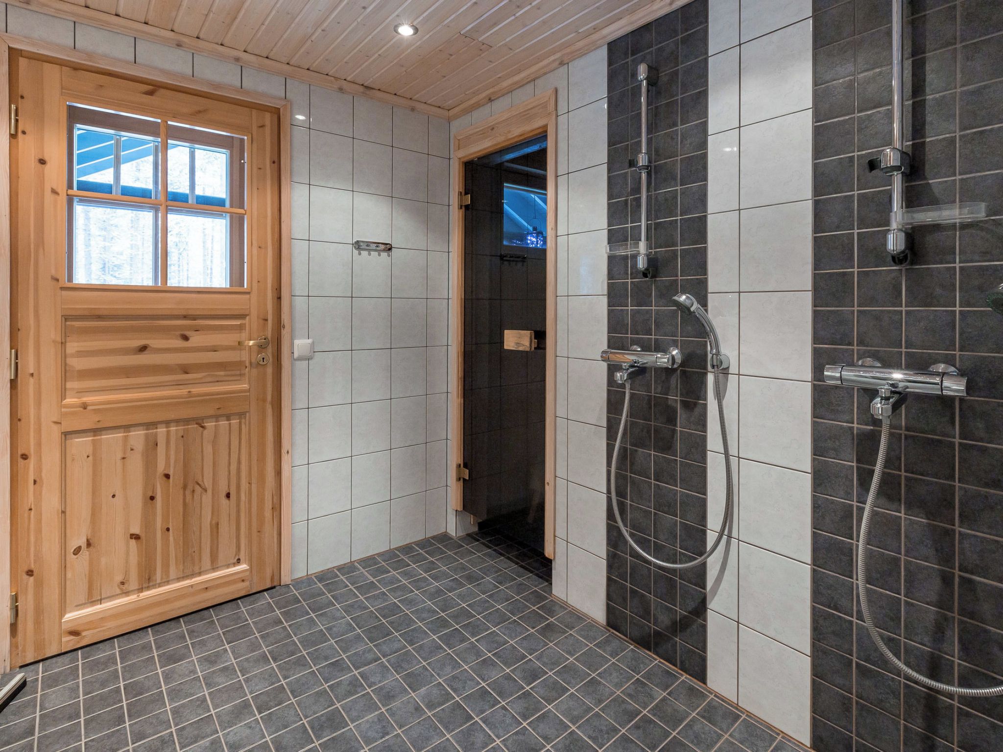 Un bagno con porta in legno, pareti piastrellate e zona doccia dotata di diversi rubinetti.