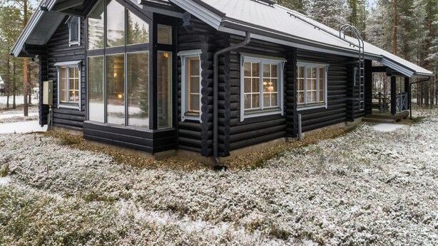 A log cabin with large windows and a sloped roof, surrounded by snow-covered ground and trees.