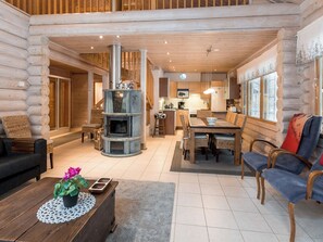 Het interieur van een blokhut, met een houtkachel in het midden, een eethoek en een woonkamer met een bank en salontafel.