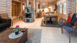 A log cabin interior with a central wood stove, a dining area, and a kitchen.