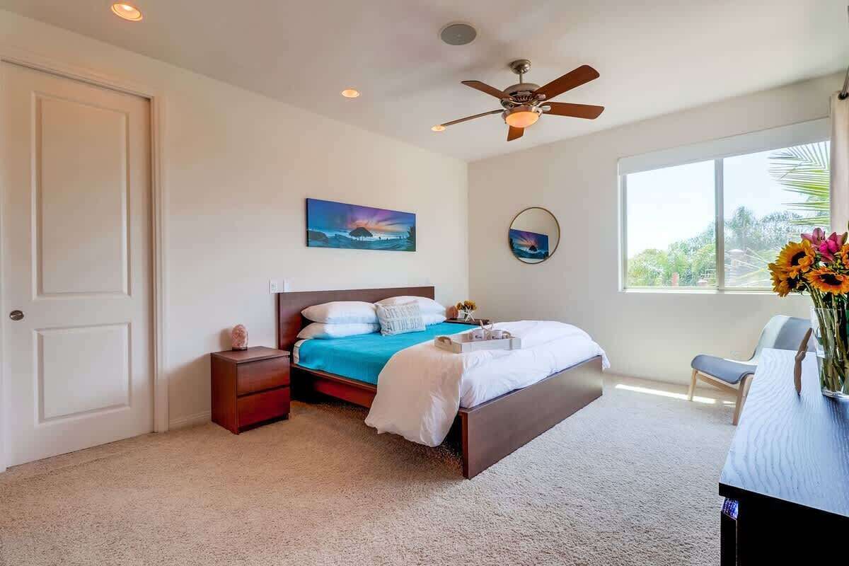 A bedroom with a bed, bedside table, nightstand, ceiling fan, artwork, window, and a desk with a vase.