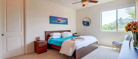 A bedroom with a bed, bedside table, nightstand, ceiling fan, artwork, window, and a desk with a vase.