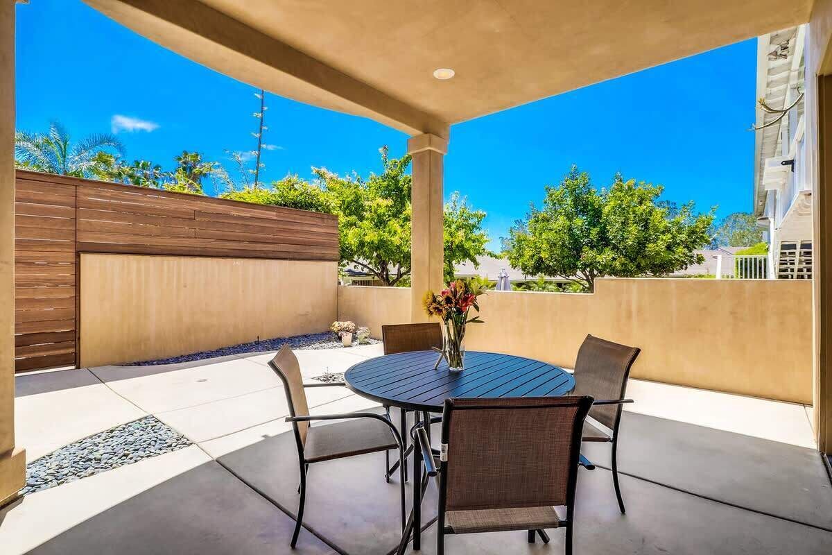 A covered patio with a round table and four chairs, a vase with flowers, and a view of trees and buildings.