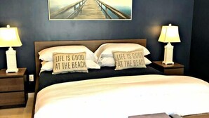 Un dormitorio con cama, dos lámparas de noche, un banco y una pared con un cuadro y almohadas con texto.