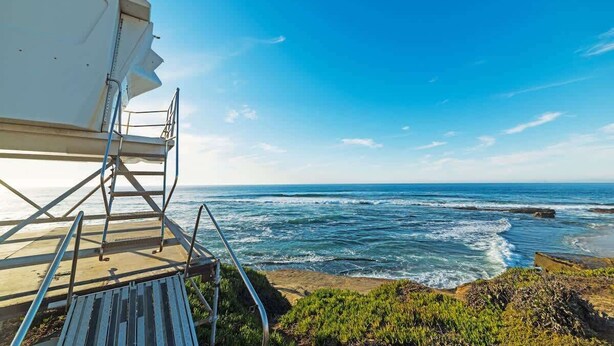 Vista costera con cielo despejado, costa rocosa y un barco blanco anclado en una plataforma de concreto.