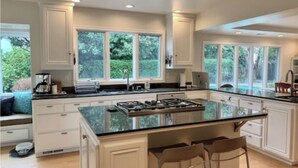 Eine geräumige Küche mit einer großen Kochinsel, weißen Schränken und großen Fenstern mit Blick auf Grünflächen.