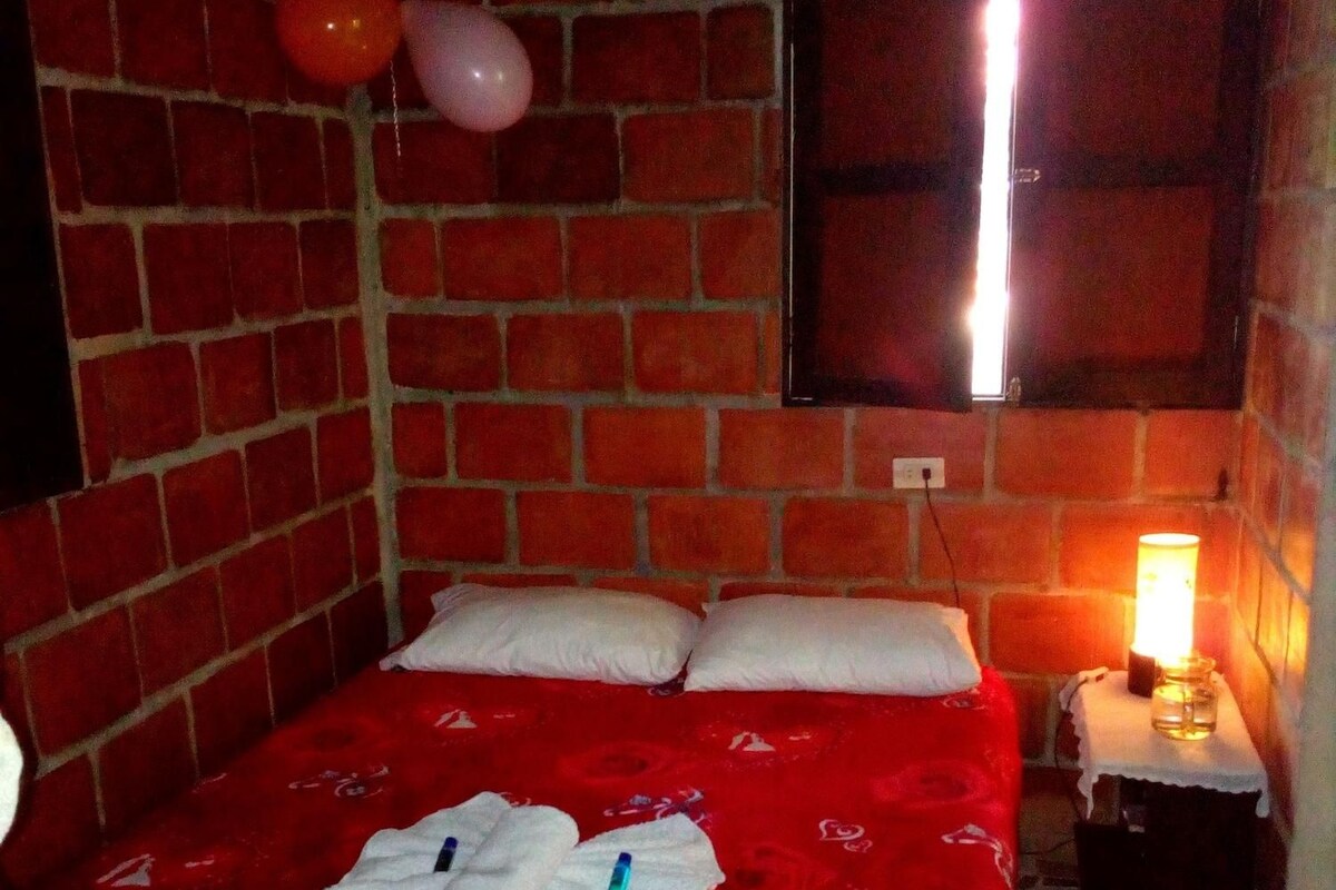 A room with a brick wall, a bed with a red bedspread, white pillows, and a bedside table with a lamp.