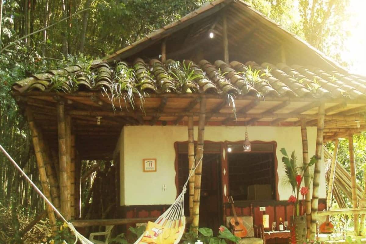 Una casa de madera rústica con techo de tejas, una hamaca y una pequeña mesa con una tetera.
